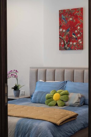 Un dormitorio con cama, almohadas, una colcha, un cuadro en la pared y una planta en maceta.