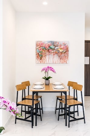 Un comedor con una mesa dispuesta para cuatro personas, rodeada de sillas de madera con estructura negra. Sobre la mesa cuelga un cuadro grande con motivos florales.
