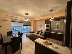 Ein modernes Wohnzimmer mit einer Couch, einem Esstisch und Stühlen, einer Küche mit Geräten sowie einem großen Fenster mit Blick.