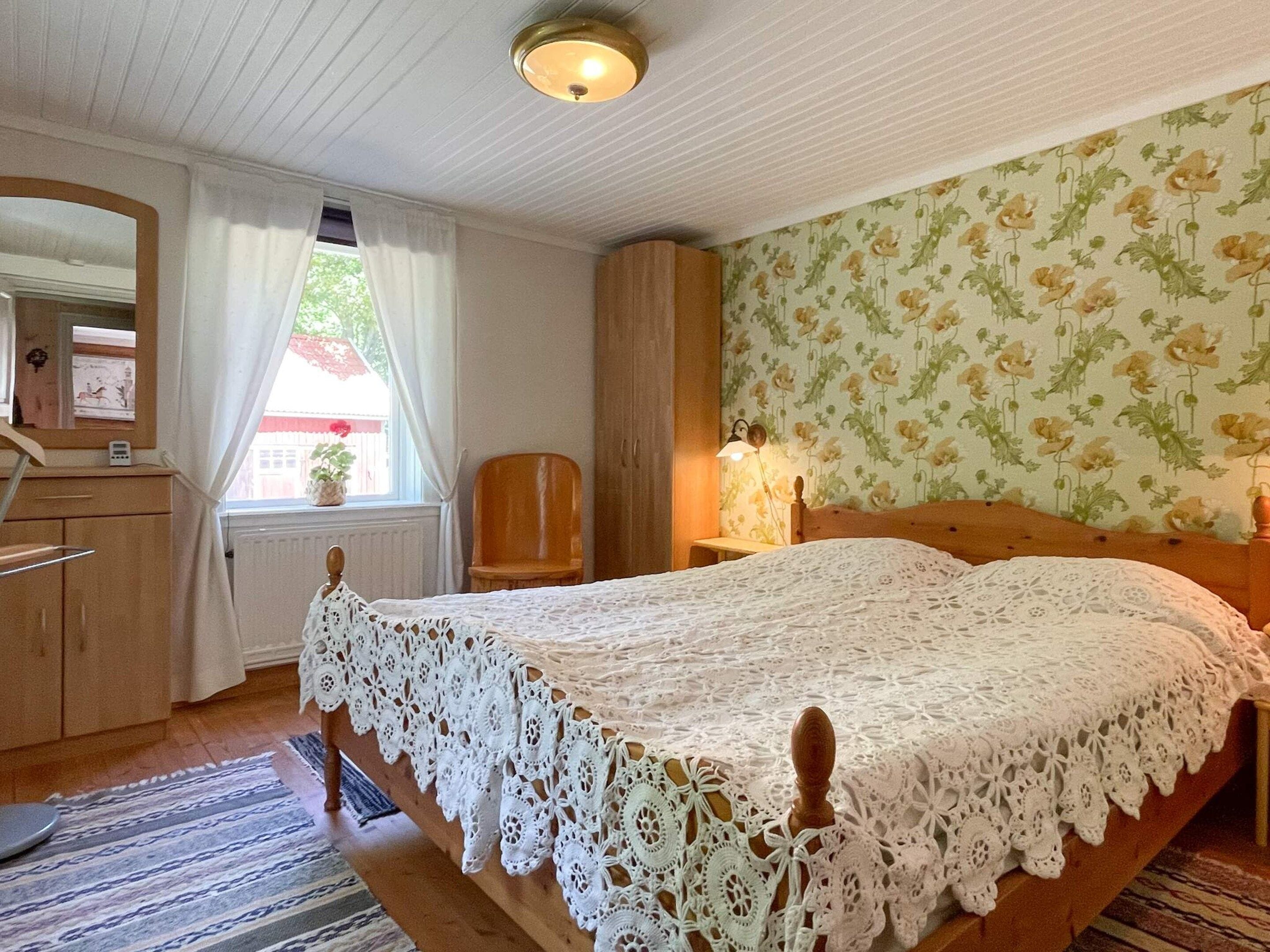Ein Schlafzimmer mit einem hölzernen Bett, einem Nachttisch, einem Sessel, einer Kommode und einem Fenster mit Vorhängen.