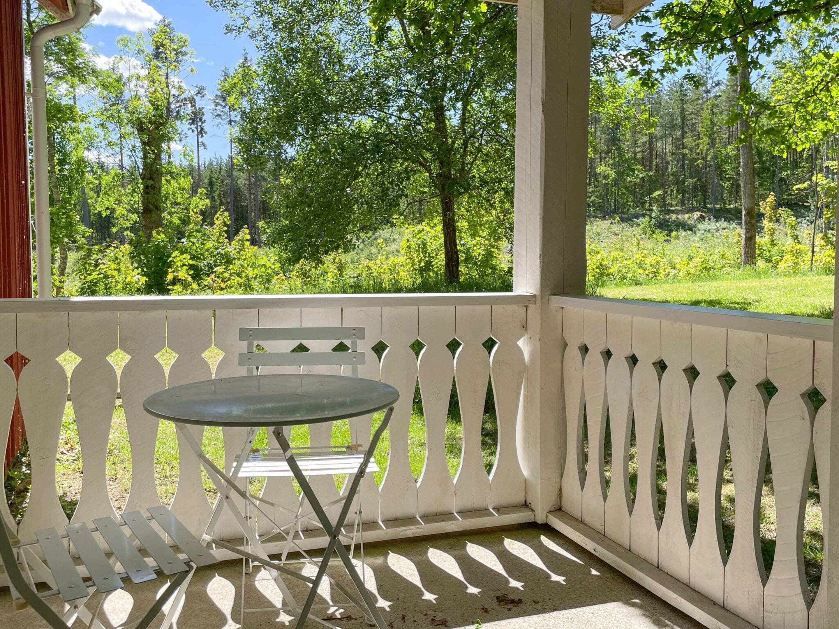 A covered outdoor area with a table and chairs, surrounded by a white fence with decorative cutouts, overlooking a forested area.