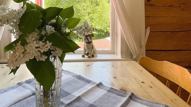 Une table en bois ornée d’un vase de fleurs blanches, recouverte d’une nappe à carreaux, et offrant une vue sur un arbre à travers une fenêtre.