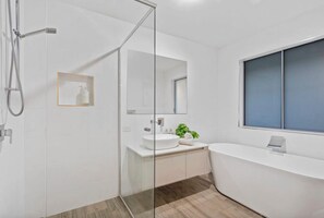 A modern bathroom with a glass shower enclosure, a white sink, a large bathtub, and a window with blinds. - The perfect family home 400mtrs from Burleigh Beach & 300mtrs from James Street. (Burleigh Waters)