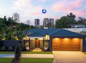 A modern house with a garage, a covered entryway, and a view of tall buildings in the background. - The perfect family home 400mtrs from Burleigh Beach & 300mtrs from James Street. (Burleigh Waters)