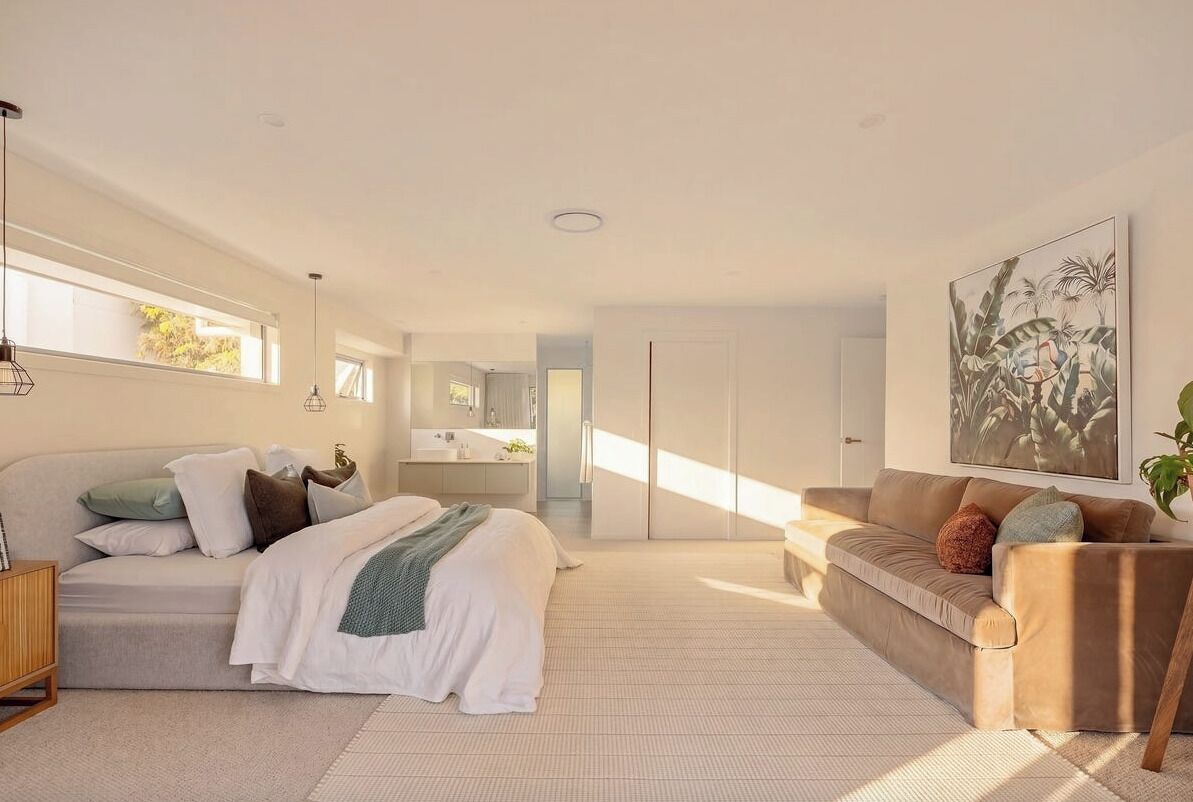 A modern bedroom with a bed, a sofa, a large window, and a bathroom in the background.