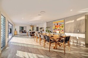 A spacious dining area with a long table and chairs, a kitchen in the background, and a large abstract painting on the wall. - The perfect family home 400mtrs from Burleigh Beach & 300mtrs from James Street. (Burleigh Waters)