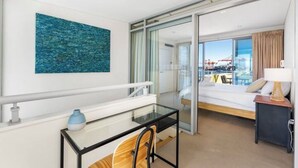 A modern hotel room with a glass partition, a bed, a desk with a chair, a lamp, and a painting on the wall. - Stylish Coastal Penthouse with Port Views (North Fremantle)