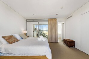 Una habitación de hotel moderna con una cama grande, un banco de madera pequeño, un balcón con vistas y una puerta corrediza de vidrio.