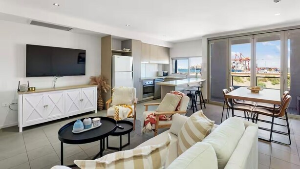 A modern living room with a sofa, coffee table, and chairs, a kitchen area, a dining table, and a large TV. - Stylish Coastal Penthouse with Port Views (North Fremantle)