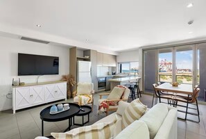 A modern living room with a sofa, coffee table, and chairs, a kitchen area, a dining table, and a large TV.