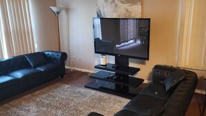 A living room with a black leather sofa, a black entertainment unit with a flat-screen TV, a standing lamp, and a potted plant. - 2 Master bedrooms condo with Two walk in Closets and half bath main floor (Indianapolis)