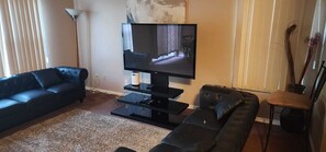 Uma sala de estar com um sofá de couro preto, um rack preto com uma TV de tela plana, um abajur e um vaso de plantas.