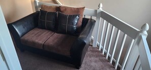 Un sofá de cuero negro con cojines marrones en un rincón de una habitación con baranda blanca y suelo alfombrado.