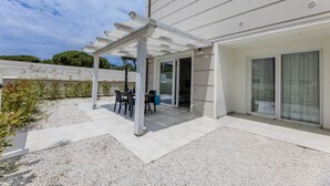 Eine überdachte Terrasse im Freien mit Tisch und Stühlen, eine Kiesfläche und ein Gebäude mit weißer Fassade und großen Fenstern.