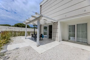 Eine überdachte Terrasse im Freien mit Tisch und Stühlen, eine Kiesfläche und ein Gebäude mit weißer Fassade und großen Fenstern.