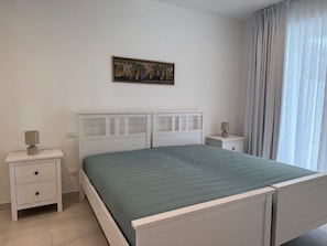 A bedroom with a bed, two bedside tables, a nightstand, a wall-mounted painting, and a window with curtains. - Appartamento Montecristo - Montecristo (Capoliveri)