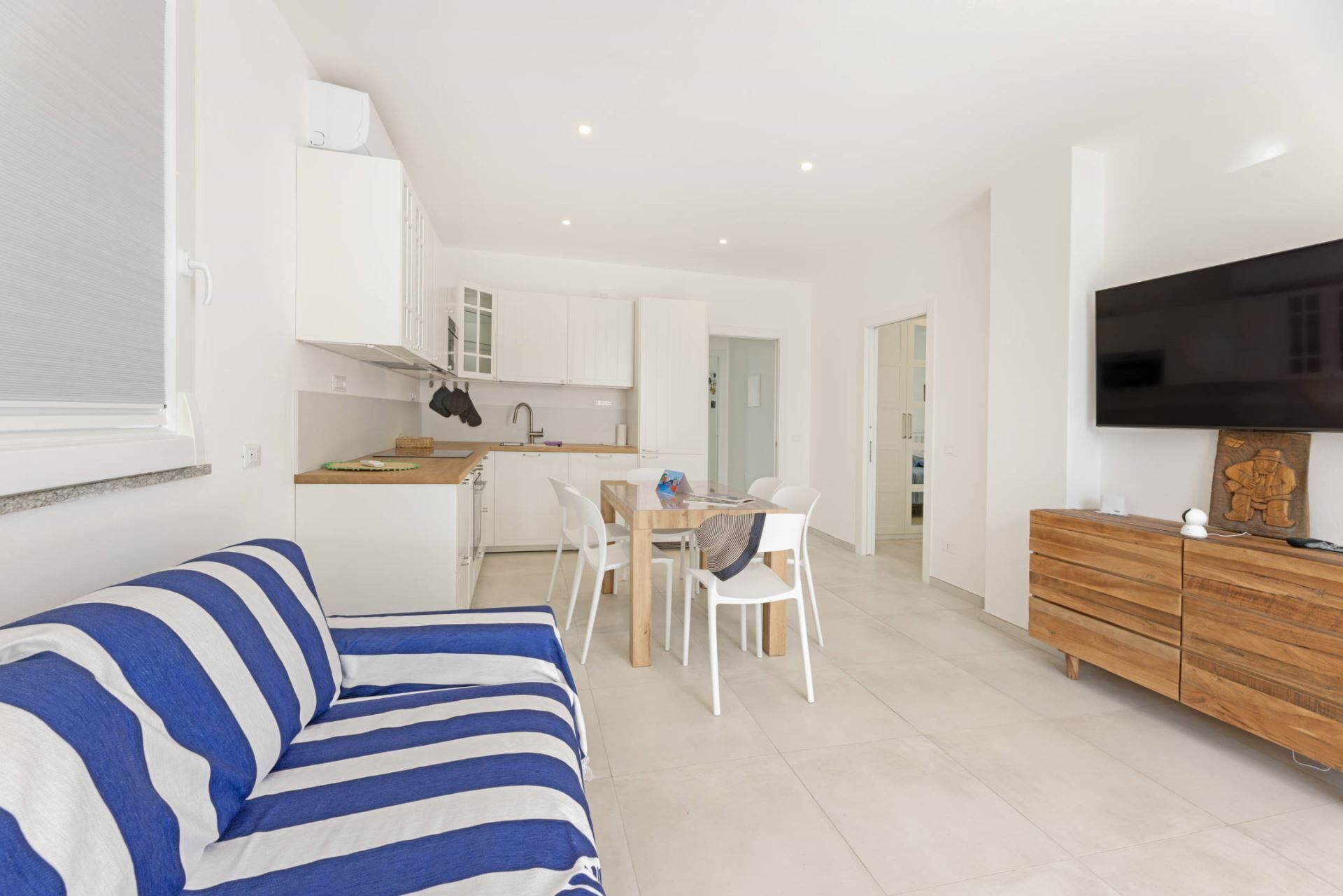 A modern living space with a striped sofa, a dining area with white chairs, a kitchen with white cabinets, and a TV on the wall.