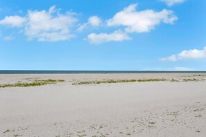 Ein Strand mit Sand und spärlicher Vegetation, ein klarer Himmel und ein weiter Horizont.