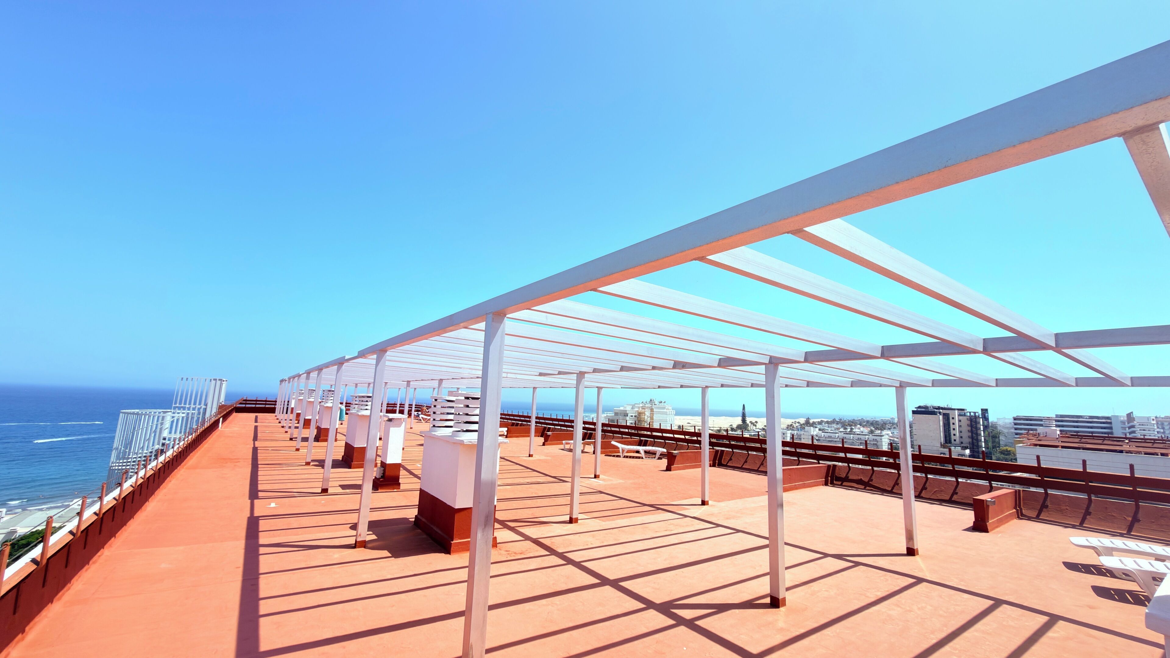 Una terraza en la azotea con vistas despejadas al océano y al paisaje urbano, con pérgolas blancas y tumbonas.