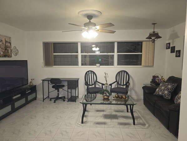A living room with a TV, a desk, two chairs, a coffee table, a sofa, a ceiling fan, and a window with curtains.