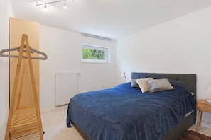 Un dormitorio con cama, perchero de madera, radiador, ventana y mesita de noche.
