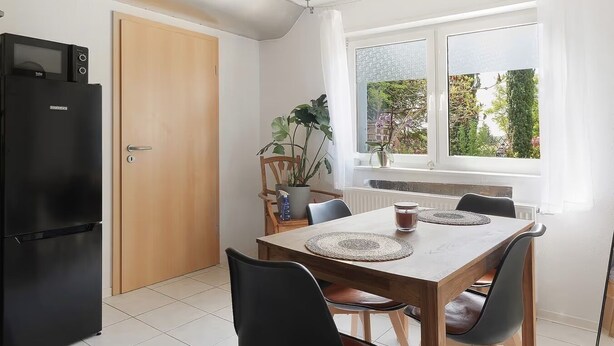 A kitchen with a black refrigerator, wooden table, chairs, a potted plant, and a window with a view of greenery.