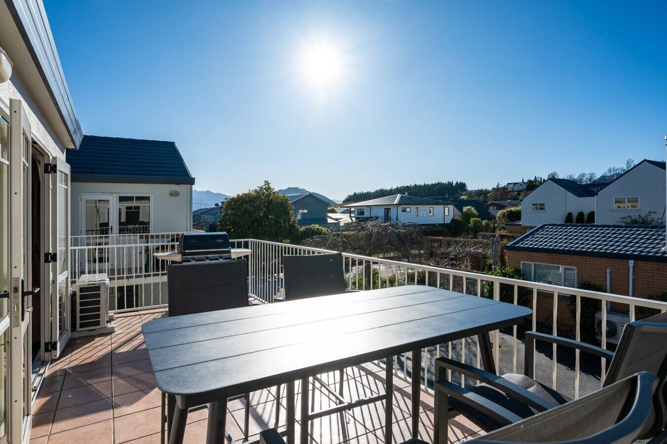A balcony with a table and chairs, a grill, and a view of houses and mountains.
