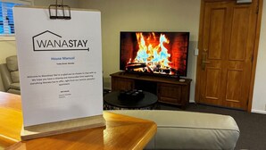 A room with a television displaying a fireplace, a wooden table, a couch, and a door. - Central Wanaka 3BR Apartment – Walk to Lake & Town (Wānaka)