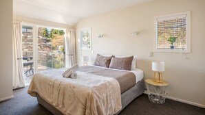 A bedroom with a bed, a bedside table with a lamp, a window with a plant, and a balcony with curtains. - Central Wanaka 3BR Apartment – Walk to Lake & Town (Wānaka)
