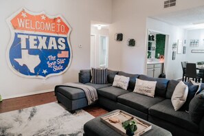 Ein Wohnzimmer mit einem Ecksofa, einem Couchtisch und einem Wandschild mit der Aufschrift 'WELCOME TO TEXAS The Lone Star State'.