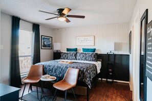Ein Schlafzimmer mit einem Bett, zwei Stühlen, einem Schreibtisch, einem Deckenventilator und einem Fenster mit Vorhängen.
