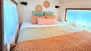 A cozy room with a wooden ceiling, a bed with a quilted bedspread, two woven wall hangings, and a window with sheer curtains.