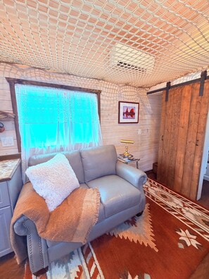 A cozy room with a grey sofa, a patterned rug, a wooden door, and a window with sheer curtains.