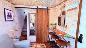 A cozy room with a wooden ceiling, a sofa, a dining area with stools, and a staircase leading to another room.
