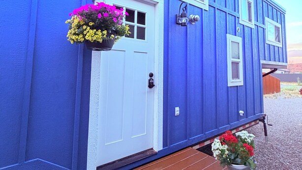 A blue mobile home with a white door, windows, and a small porch area with flower planters.