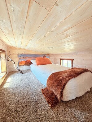 A cozy cabin bedroom with a bed, a window, a wooden ceiling, and a view of the sky.