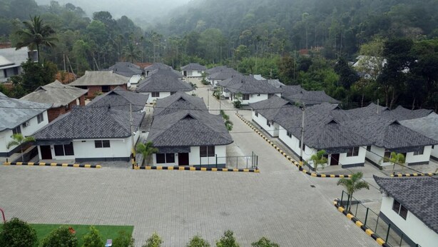 A residential area with multiple houses, some with sloped roofs, surrounded by greenery and hills.