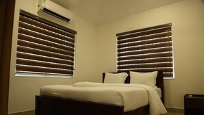 A bedroom with a bed, two windows with blinds, an air conditioning unit, and a nightstand.