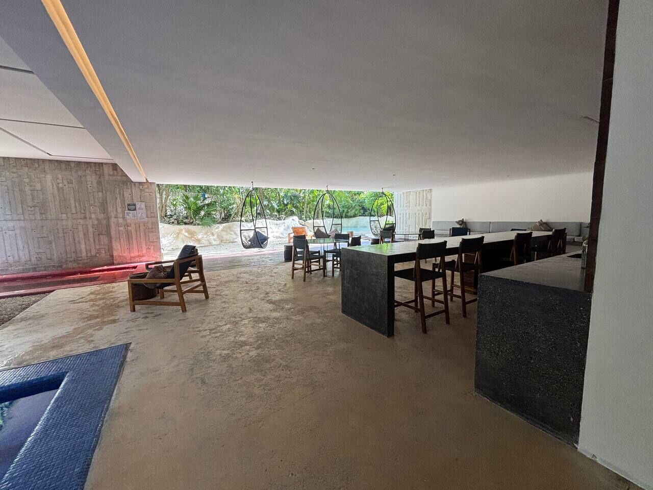 Un espacio interior amplio con una mesa y sillas de comedor, una pequeña zona de estar con una silla, y vistas a zonas verdes.