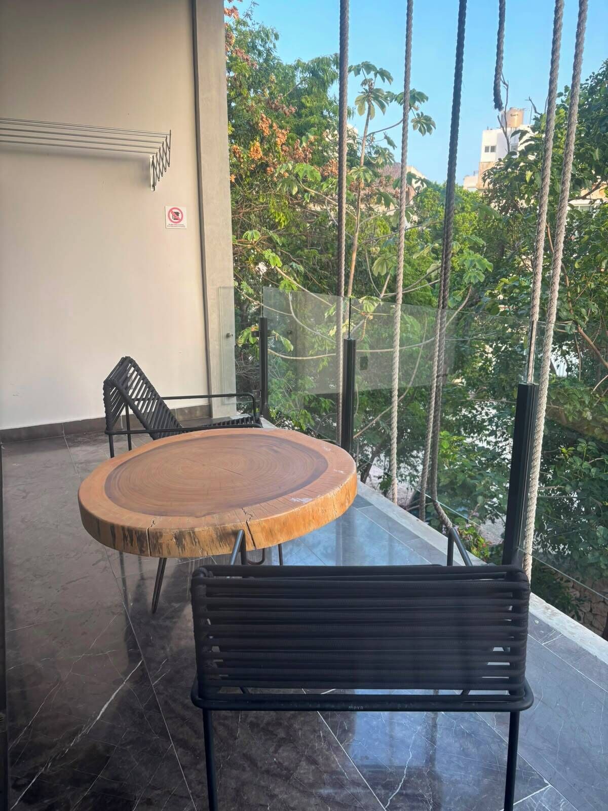 A balcony with a round wooden table and a black bench, surrounded by glass railings and trees.