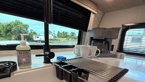 Uma cozinha com pia, cafeteira e janela com vista para árvores e uma caravana.