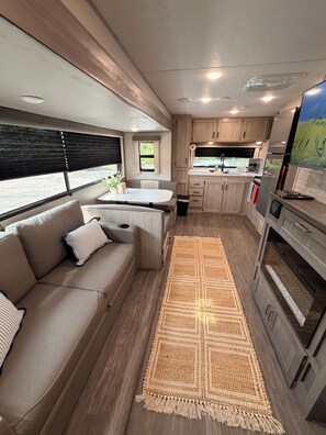 A compact RV interior with a kitchenette, a seating area with a sofa, and a large window. -  Quiet Texas Retreat  New 2026 RV with Full Amenities (Springtown)