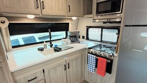Cozinha compacta de um veículo recreativo, com armários de madeira, micro-ondas, fogão e geladeira.
