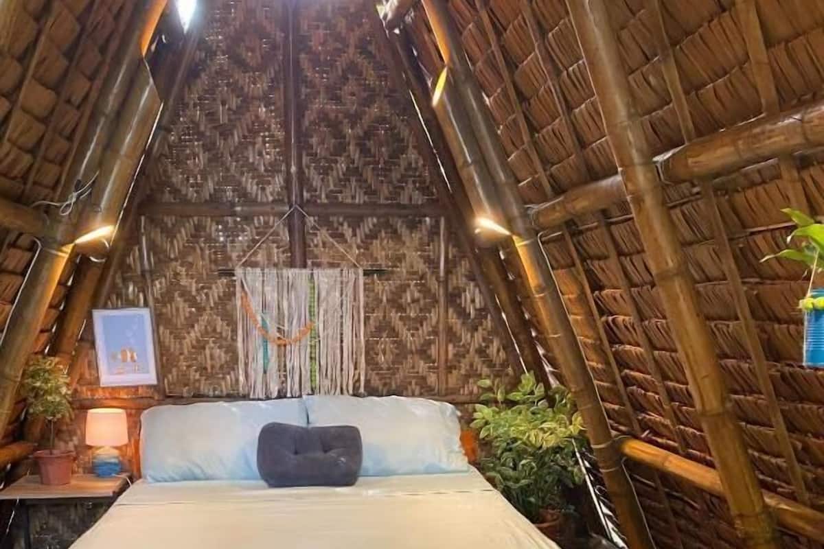 A room with a bed, a wooden floor, a thatched roof, and a wall with a woven pattern.