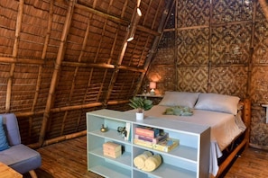 Ein Zimmer mit Strohdach, einem Bett, einem Bücherregal und einem kleinen Tisch mit einer Pflanze.