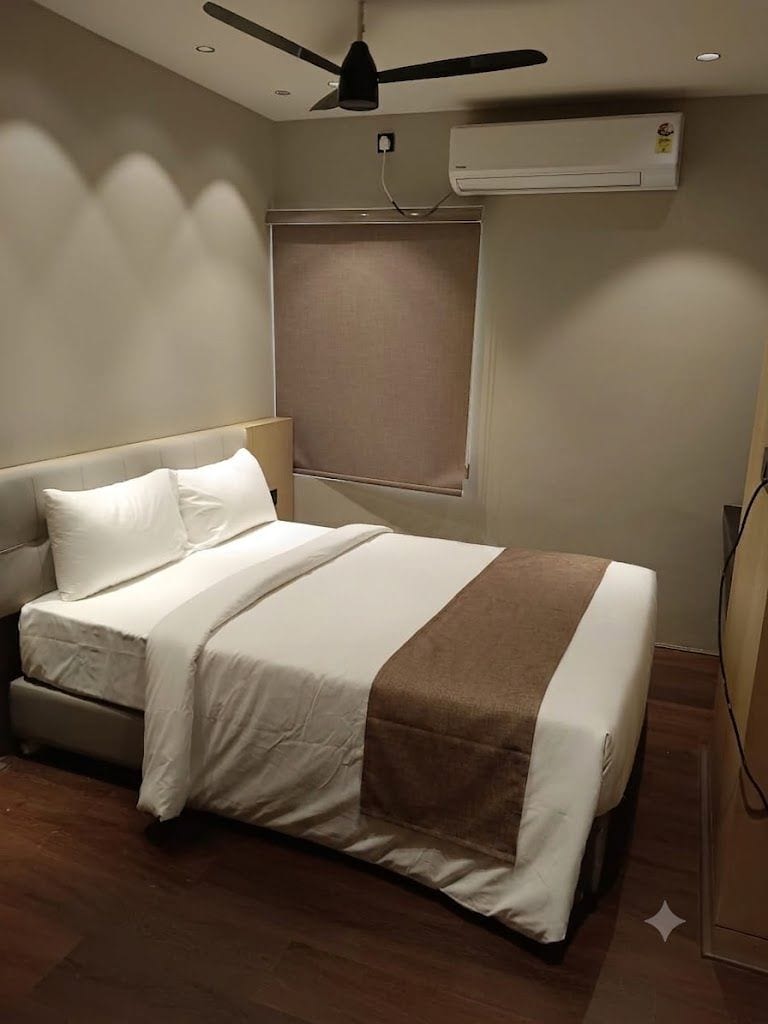 Habitación de hotel con cama, ventilador de techo, equipo de aire acondicionado y ventana con persiana.
