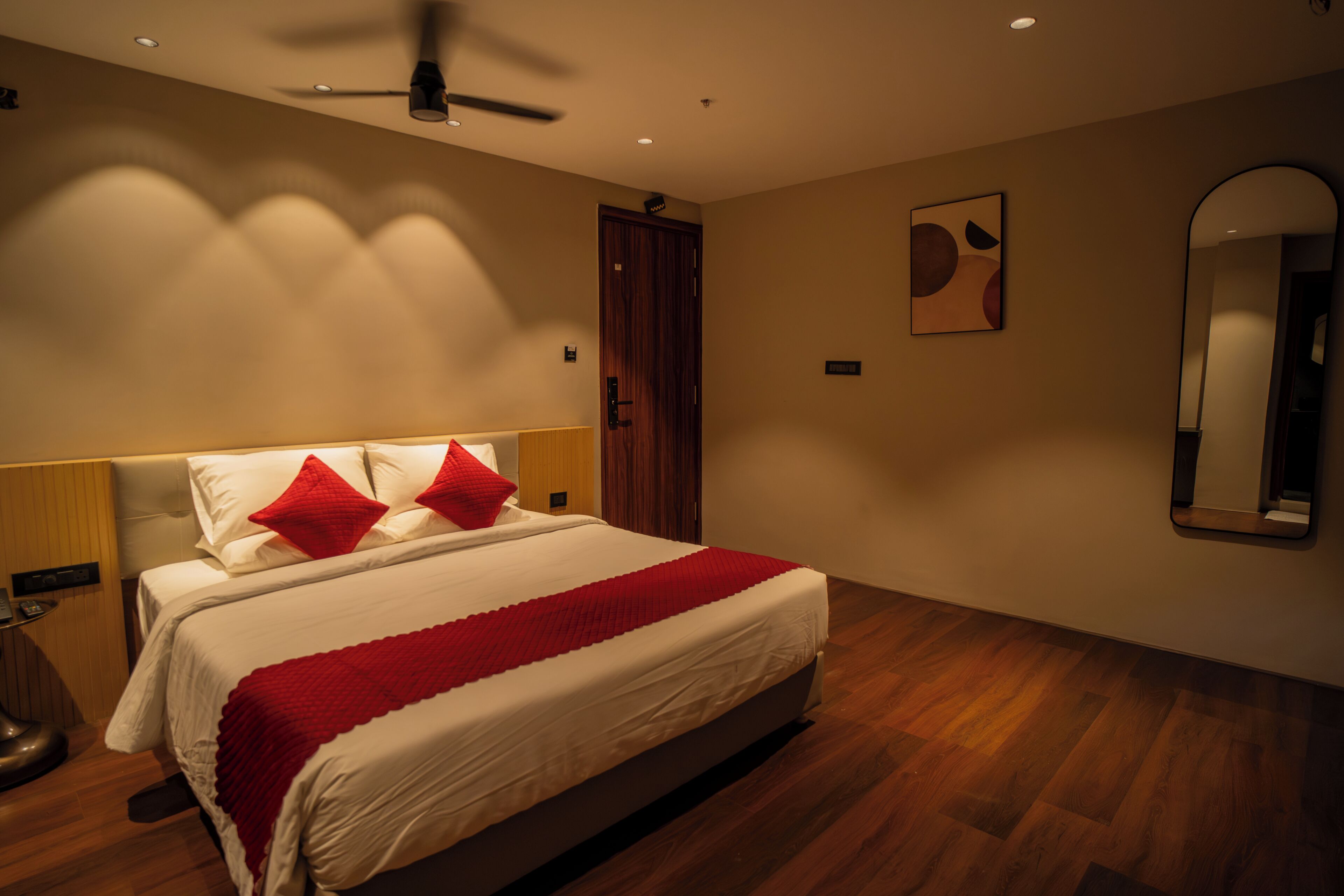 A bedroom with a bed, a ceiling fan, a wooden door, a wall-mounted mirror, and a red pillow.