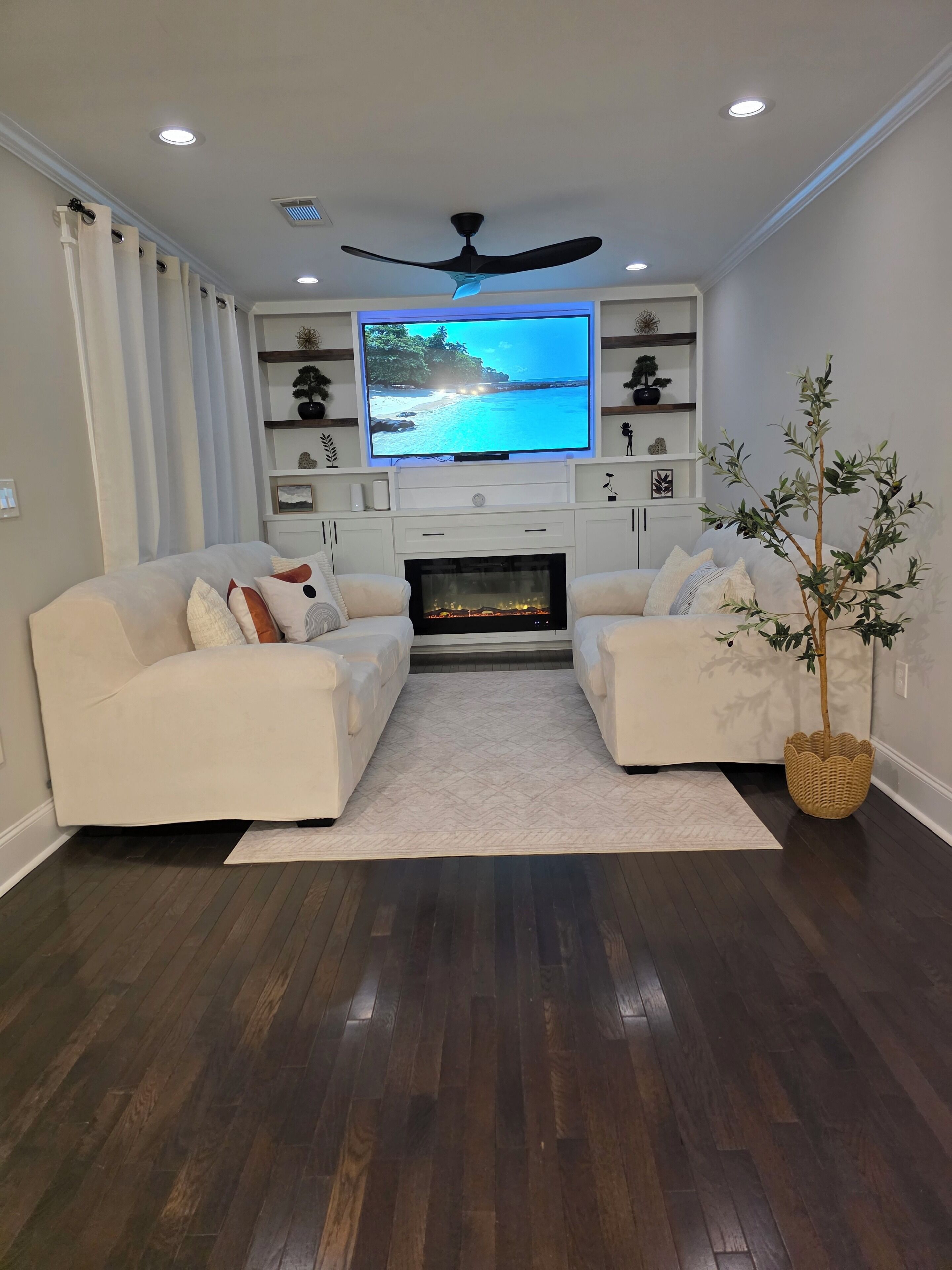 A living room with a fireplace, two white sofas, a television, and a ceiling fan.