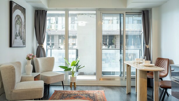 Ein modernes Hotelzimmer mit einer großen Schiebeglaswand, einem Esstisch, zwei Sesseln, einem kleinen Beistelltisch mit Blumenvase und einer Zimmerpflanze.