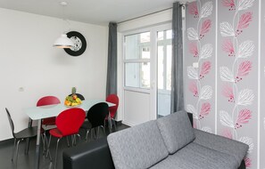 Ein modernes Zimmer mit Esstisch, roten Stühlen, einem grauen Sofa, einer Hängelampe und einem Fenster mit Vorhängen.